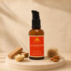 An amber glass face moisturizer bottle with a black pump and a red-orange label reading 'AMHARAWA NATURAL face moisturizer with Sandalwood' is displayed on a round stone pedestal with small props including a gold bowl, wooden pieces, and almonds against a textured beige background.