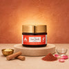A jar of Calypso Organic Face Mask with Shea Butter sits on a round stone pedestal, surrounded by cosmetic ingredients including a small bowl of white scrub, cinnamon sticks, a pile of brown powder, and pink petals against a textured terracotta background.