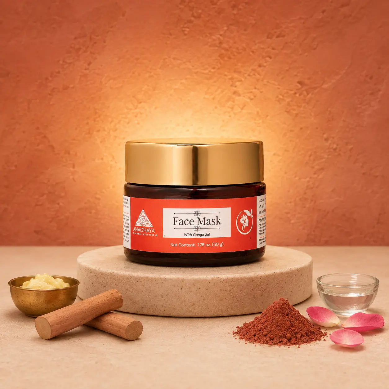 A jar of Calypso Organic Face Mask with Shea Butter sits on a round stone pedestal, surrounded by cosmetic ingredients including a small bowl of white scrub, cinnamon sticks, a pile of brown powder, and pink petals against a textured terracotta background.