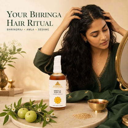A woman with long, dark, shiny hair looks down at a bottle of Bhringraha Hair Oil placed on a marble tray beside green bhringra leaves, sesame seeds, and a brass bowl. The scene is warmly lit with decorative plants and text reading 'YOUR BHRINGA HAIR RITUAL' and 'BHRINGRAHA + AMLA + SESAME'.